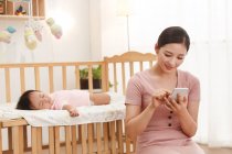 Sonriente joven asiático mujer usando smartphone mientras bebé durmiendo en cuna - foto de stock