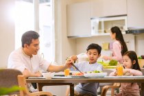 Padre con figli seduto e mangiare a tavola, madre che cucina in cucina — Foto stock