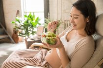 Vue latérale d'une jeune femme enceinte souriante qui mange une salade de légumes saine à la maison — Photo de stock