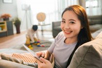 Heureux jeune asiatique femme à l'aide numérique tablette et sourire à la caméra tandis que fils jouer avec jouets derrière à la maison — Photo de stock