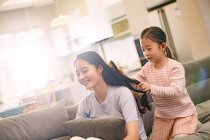 Adorable little daughter combing hair to smiling young mother sitting on couch — Stock Photo