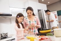 Счастливая мать и дочь готовят вместе, пока отец открывает холодильник на кухне — стоковое фото