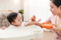 Schnappschuss von junger Mutter, die Schüssel mit Löffel in der Hand hält und adoptierbares Säugling zu Hause füttert — Stockfoto