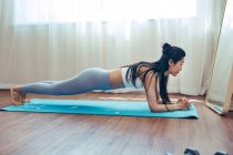 Vista lateral de la joven deportista practicando la posición de tablón en la esterilla de yoga en casa - foto de stock