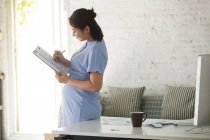 Side view of young pregnant businesswoman writing on clipboard — Stock Photo