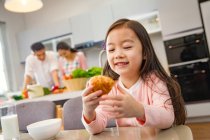 Adorabile bambino felice che tiene il croissant mentre i genitori cucinano dietro in cucina — Foto stock