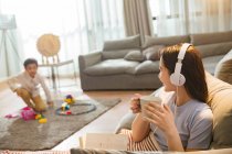 Giovane donna in cuffie in possesso di tazza e guardando il figlio che gioca con i giocattoli su tappeto — Foto stock