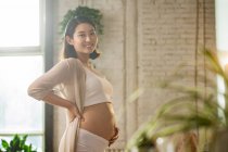 Vista laterale di allegra giovane donna asiatica incinta toccare la pancia e sorridere alla fotocamera, messa a fuoco selettiva — Foto stock