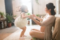 Вид сбоку на беременную молодую мать и симпатичную маленькую дочку, держащуюся за руки и играющую дома — стоковое фото