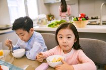 Adorable asian brother and sister having breakfast together while mother cooking behind in kitchen — Stock Photo