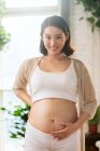Feliz joven embarazada asiático mujer tocando vientre y sonriendo en cámara - foto de stock
