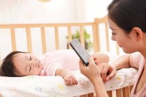 Schnappschuss von junger Mutter mit Smartphone, während ihr entzückendes Baby im Kinderbett schläft — Stockfoto