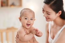 Glückliche junge Mutter trägt entzückend lächelndes Baby zu Hause — Stockfoto
