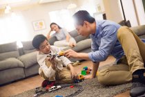 Счастливый азиатский отец и сын играют с игрушками на ковре, мать сидит на диване позади — стоковое фото