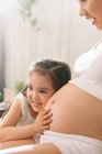Entzückende glückliche kleine Mädchen umarmt und hört Bauch der schwangeren Mutter zu Hause, abgeschnittenen Schuss — Stockfoto