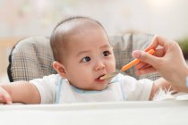 Cropped shot of mother holding spoon and feeding adorable infant at home — Stock Photo
