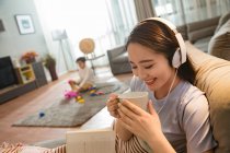 Sonriente mujer joven en auriculares sentados en el sofá y sosteniendo la taza mientras el hijo juega con juguetes detrás en casa - foto de stock