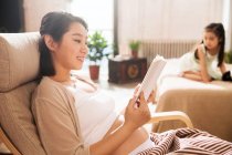 Vista lateral de la madre embarazada sonriente leyendo libro e hija pequeña jugando detrás en casa - foto de stock