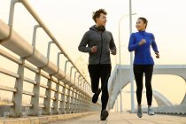 Счастливая спортивная молодая азиатская пара улыбается друг другу и бегает вместе по мосту — стоковое фото
