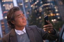 Serious mature asian man leaning at car and using smartphone in night city — Stock Photo
