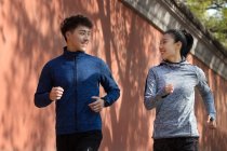 Atlética joven pareja en ropa deportiva sonriendo entre sí y corriendo juntos en la calle - foto de stock
