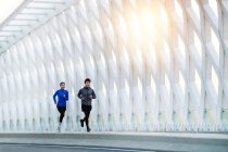 Sonriente joven asiático macho y hembra atletas en sportswear jogging juntos en moderno puente - foto de stock