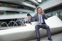 Красивый азиатский бизнесмен, использующий ноутбук и разговаривающий на смартфоне возле фонтана в современном бизнес-центре — стоковое фото