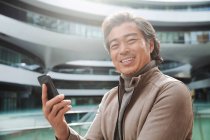 Веселий зрілий азіатських чоловік тримає смартфон і посміхається на камеру — стокове фото