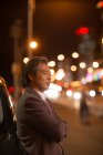 Side view of pensive mature asian man standing with crossed arms beside car in night city — Stock Photo
