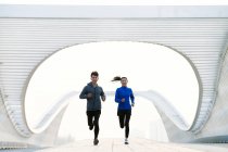 Front view of smiling young male and female athletes jogging together on modern bridge — Stock Photo