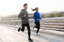 Smiling young asian man and woman running together on bridge — Stock Photo