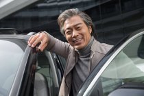 Guapo feliz asiático hombre de pie cerca de coche y sonriendo a la cámara - foto de stock