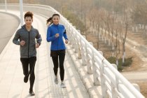 Высокий угол обзора спортивной молодой азиатской пары, бегущей по мосту и улыбающейся перед камерой — стоковое фото