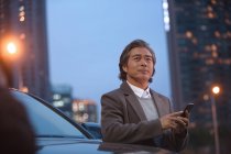 Зрілий азіатський чоловік стоїть біля машини і використовує смартфон вночі — стокове фото