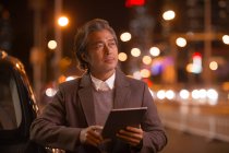 Reifer asiatischer Mann benutzt digitales Tablet neben Auto und schaut weg — Stockfoto