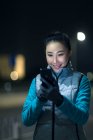 Улыбающаяся молодая азиатка в спортивной одежде с помощью смартфона в ночном городе — стоковое фото
