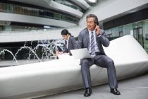 Handsome asian businessman using laptop and talking by smartphone near fountain in modern business center — Stock Photo
