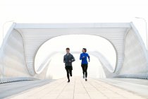 Вид спереди улыбающихся молодых азиатских бегунов в спортивной форме, тренирующихся вместе на мосту — стоковое фото