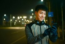 Lächelnder asiatischer Mann mit Kopfhörern und Sportbekleidung mit Smartphone in der nächtlichen Stadt — Stockfoto