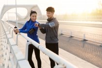 Lächelndes junges Paar überprüft Armbanduhr, während es nach dem Training zusammen auf der Brücke steht — Stockfoto