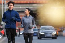 Smiling young asian sportswoman looking at boyfriend while running together on street — Stock Photo