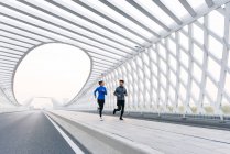 Sporty young couple looking at each other and running together on bridge — Stock Photo