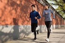 Vista completa de la joven pareja asiática en ropa deportiva sonriendo y corriendo juntos en la calle - foto de stock