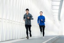 Vista completa de sonriente joven asiático hombre y mujer en ropa deportiva corriendo juntos en puente - foto de stock