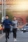 Счастливая спортивная молодая азиатская пара, улыбающаяся в камеру и бегающая вместе по улице — стоковое фото