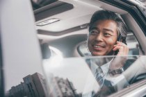 Улыбающийся азиатский бизнесмен, сидящий в машине и разговаривающий по смартфону, избирательный фокус — стоковое фото