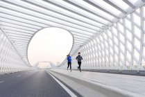 Full length view of young male and female joggers in sportswear running together on bridge — Stock Photo