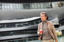Усміхнений азіатський бізнесмен тримає паперовий стаканчик і ходить біля офісної будівлі — стокове фото