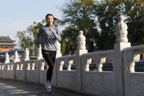 Longitud completa vista de hermosa sonriente asiático chica en sportswear corriendo al aire libre - foto de stock