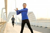 Junge Frau streckt sich und lächelt in die Kamera, während sportlicher Mann auf Brücke hinterherläuft — Stockfoto